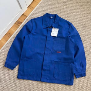 May include: A vibrant blue work jacket featuring a button-down design, two chest pockets, and two lower pockets. The jacket has long sleeves and a collar. A tag is attached to the chest pocket. A small red logo is visible on the lower pocket.