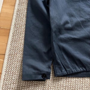 Puede incluir: Primer plano de una manga de chaqueta azul marino con un bot&oacute;n negro. La tela tiene un aspecto ligeramente texturizado. La chaqueta est&aacute; sobre una alfombra beige y marr&oacute;n estampada.
