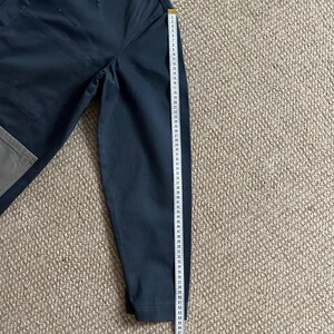 Puede incluir: Pantalones azul marino oscuro con un bolsillo gris y una cinta m&eacute;trica amarilla. La cinta muestra medidas de hasta 170 cm. Los pantalones est&aacute;n sobre una superficie texturizada de color marr&oacute;n claro.