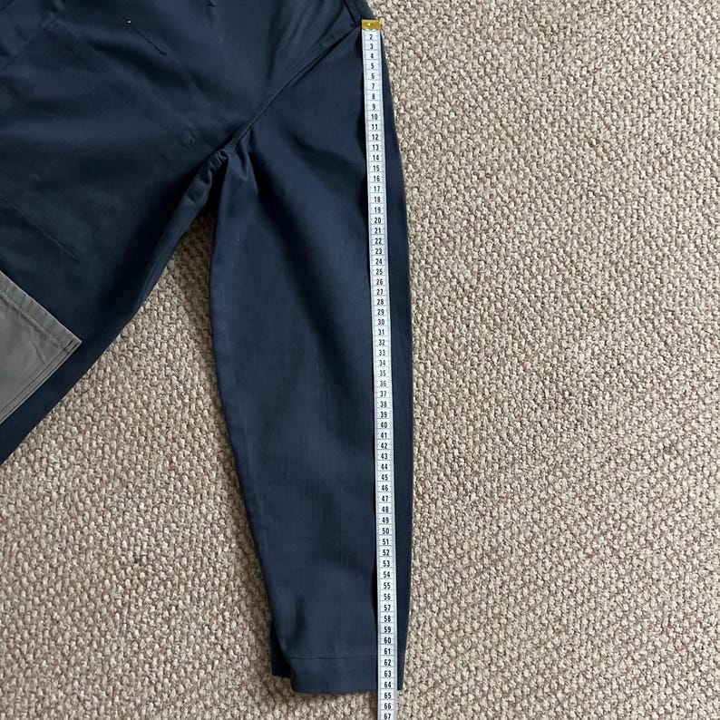 Puede incluir: Pantalones azul marino oscuro con un bolsillo gris y una cinta m&eacute;trica amarilla. La cinta muestra medidas de hasta 170 cm. Los pantalones est&aacute;n sobre una superficie texturizada de color marr&oacute;n claro.