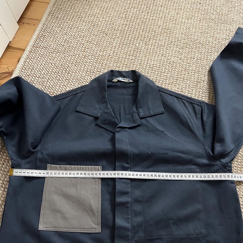 Puede incluir: Una camisa de trabajo azul marino con un bolsillo gris y un cuello. Una cinta m&eacute;trica se coloca sobre la camisa, mostrando una longitud de aproximadamente 61 cm. La camisa tiene mangas largas y cierre de botones.