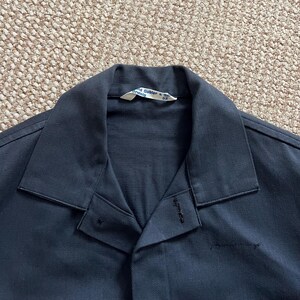Puede incluir: Primer plano de una camisa de trabajo azul marino con cuello y cierre de botones. La camisa tiene una etiqueta en el cuello con el texto "David Gabler & Co. Drehtig". La tela parece ser un material tejido duradero.