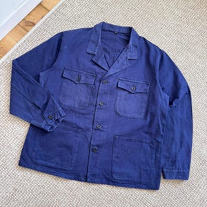 May include: A long-sleeved navy blue work jacket featuring a collared neckline, a button-down front, two chest pockets with buttoned flaps, and two large lower patch pockets. The garment is constructed with visible stitching and dark buttons.