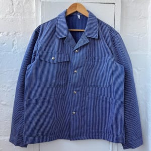May include: Blue and white striped work jacket with four front pockets and gold-colored buttons. The jacket is hanging on a wooden hanger against a white background. The jacket has a classic, utilitarian design.