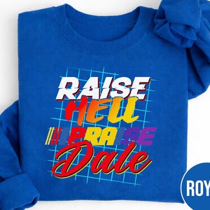 May include: Royal blue sweatshirt with the text "RAISE HELL PRAISE Dale" in a retro font. The text is in white, red, yellow, and purple. The design is on a grid background. The sweatshirt has long sleeves.