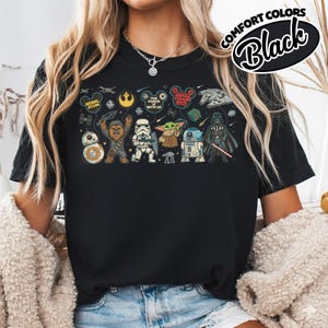 Puede incluir: Camiseta negra con un dise&ntilde;o de Star Wars. El gr&aacute;fico incluye personajes como Darth Vader, Chewbacca y Baby Yoda, junto con las frases "The Force is with Us" y "Empire Strikes Back". La camiseta tambi&eacute;n tiene el texto "Comfort Colors Black".