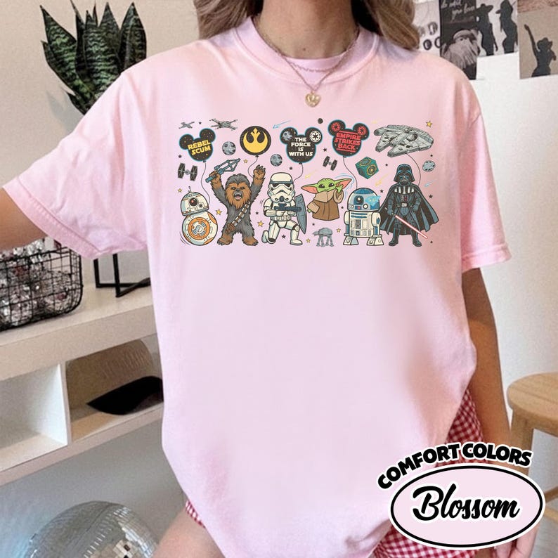 Puede incluir: Camiseta rosa claro Comfort Colors con un dise&ntilde;o de Star Wars. El gr&aacute;fico presenta personajes como Darth Vader, Chewbacca y Baby Yoda, junto con frases. La camiseta tiene la palabra "Blossom" en un &oacute;valo blanco.
