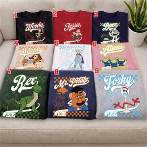 Pode incluir: Nove camisetas coloridas com personagens do filme de anima&ccedil;&atilde;o Toy Story. Cada camisa exibe o nome e a imagem de um personagem, incluindo Woody, Jessie, Buzz, Slinky, Bo Peep, Aliens, Rex, Mr. Potato Head e Forky. O ano de 1995 tamb&eacute;m est&aacute; impresso em cada camisa.