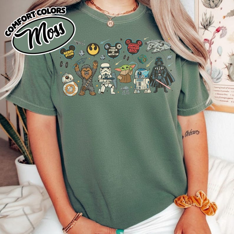 Puede incluir: Camiseta verde musgo con personajes y logotipos de Star Wars. El dise&ntilde;o incluye a BB-8, Chewbacca, un Stormtrooper, Grogu, R2-D2 y Darth Vader. La camiseta tambi&eacute;n tiene el texto "COMFORT COLORS" y "Moss" en una fuente curva.