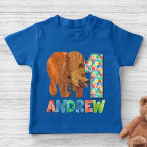 May include: A blue toddler t-shirt featuring a brown bear illustration and the number 1. The name "ANDREW" is printed in colorful letters below the bear. The bear illustration has a textured appearance.