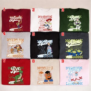 Pode incluir: Nove camisolas de gola redonda em v&aacute;rias cores, cada uma com um personagem diferente do filme de anima&ccedil;&atilde;o "Toy Story". Os designs incluem Woody, Jessie, Buzz, Slinky, Bo Peep, Aliens, Rex, Mr. Potato Head e Forky, com o ano "1995" exibido.