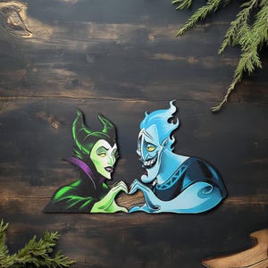 May include: A wooden cutout featuring the characters Maleficent and Hades from Disney's animated films. Maleficent is depicted in green and black, while Hades is blue. They are positioned with their hands forming a heart shape. The background is dark wood.