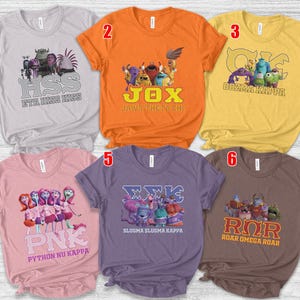 May include: A collection of six short-sleeved t-shirts in various colors, each featuring a graphic design and text. The designs include cartoon characters and Greek letter combinations. The shirts are displayed on a light-colored surface.