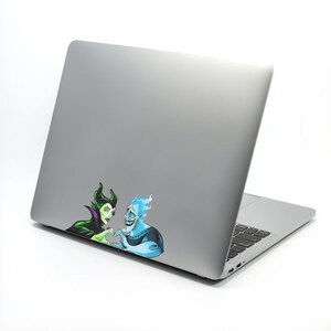 May include: A silver laptop with a sticker featuring cartoon characters Maleficent and Hades. Maleficent is green and purple, and Hades is blue. They are making a heart shape with their hands.