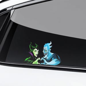 May include: A car window decal featuring cartoon characters Maleficent and Hades forming a heart shape with their hands. Maleficent is in green and purple, and Hades is blue. The decal is on a car window.