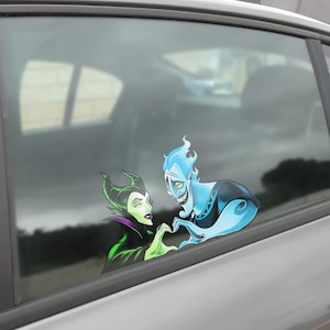 May include: A car window decal featuring cartoon characters Maleficent and Hades forming a heart shape with their hands. Maleficent is in a black and green outfit, and Hades is blue with flame-like hair. The decal is applied to a car window.