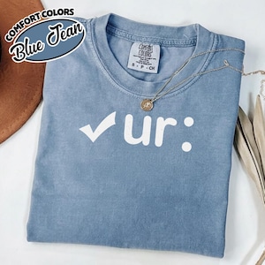 May include: A blue Comfort Colors t-shirt with the text "✓ur:" in white. The shirt has a crew neck and short sleeves. A gold necklace with a pendant is draped across the shirt. The shirt is folded on a surface.