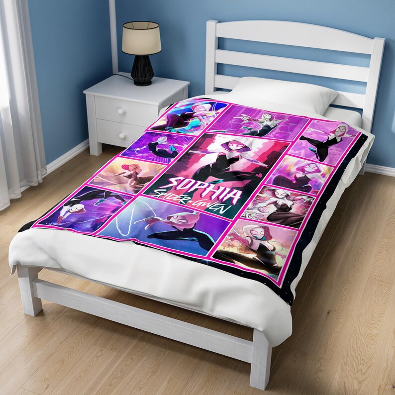 May include: A colorful blanket featuring a grid of images of a superheroine in various action poses. The blanket has a black border and the name "SOPHIA SPIDER GWEN" is prominently displayed in the center. The blanket is on a white bed.