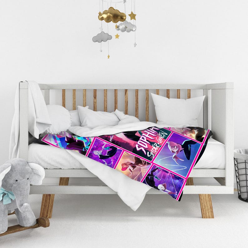 May include: A colorful blanket featuring a comic book style design with the words "Sophia Spider-Gwen". The blanket has a pink border and is draped over a white crib. A gray elephant toy and decorative hanging clouds are also visible.