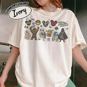 Puede incluir: Camiseta de color marfil con un gr&aacute;fico de Star Wars. El dise&ntilde;o incluye ilustraciones de dibujos animados de personajes como Chewbacca, Darth Vader y Baby Yoda, junto con elementos de texto como "Rebel Scum" y "The Force is With Us."