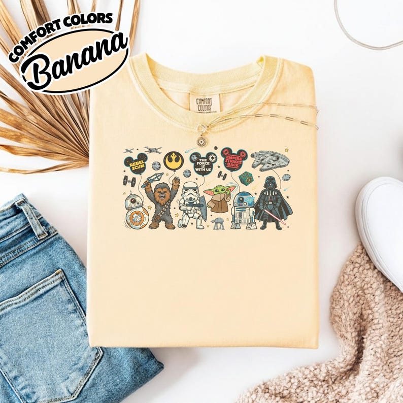 Puede incluir: Una camiseta Comfort Colors amarillo claro con un dise&ntilde;o de Star Wars. El dise&ntilde;o presenta personajes de dibujos animados como Chewbacca y Darth Vader, junto con globos tem&aacute;ticos y el texto "The Force With Us."