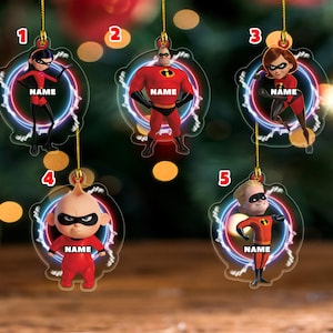 May include: Set of five acrylic Christmas ornaments featuring characters from The Incredibles. Each ornament has a character in a superhero pose, surrounded by a glowing blue and red design. Each ornament has the word "NAME" on it.