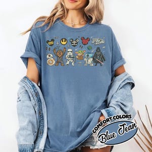 Puede incluir: Camiseta azul vaquero con un gr&aacute;fico de Star Wars. El gr&aacute;fico incluye varios personajes y elementos de la franquicia Star Wars, como Darth Vader, Chewbacca y el Halc&oacute;n Milenario. La camiseta tiene las mangas dobladas.