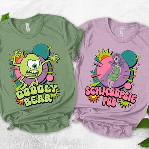 May include: Two t-shirts, one olive green and one lavender, each featuring a cartoon character design. The green shirt says "Googly Bear" and the lavender shirt says "Schmoopsie Poo" in colorful, stylized text. The designs include vibrant colors and retro graphics.