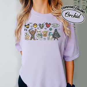 Puede incluir: Una camiseta de color lila claro con un gr&aacute;fico de personajes e iconos de Star Wars. El dise&ntilde;o incluye personajes como Chewbacca, Darth Vader y R2-D2, junto con el texto "The Force With Us". La camiseta tambi&eacute;n tiene el texto "Comfort Colors" y "Orchid".