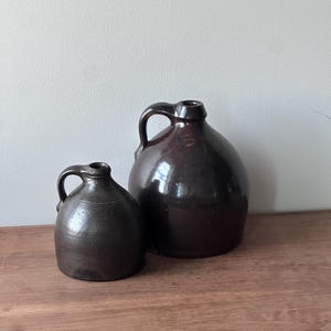 May include: Two antique brown stoneware jugs with handles. The jugs are different sizes, with a smaller one in front of a larger one. The jugs are sitting on a wooden surface.