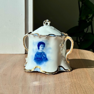 May include: White porcelain sugar bowl with a lid, featuring a blue portrait and gold trim. The handles and edges are adorned with gold accents. A decorative piece for serving sugar or other condiments.