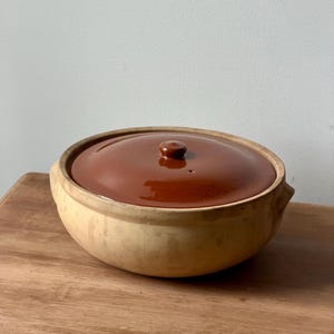 May include: A round, low-profile ceramic casserole dish with a matte, light beige exterior and a glossy, dark brown glazed lid. The lid features a small, centered knob handle, and the base has integrated side handles for easy lifting and handling.