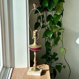 Puede incluir: Una lámpara de mesa vintage con base de latón y pantalla de cristal rojo rubí. La lámpara tiene una base de mármol y un remate decorativo. La lámpara mide aproximadamente 38 cm de altura.