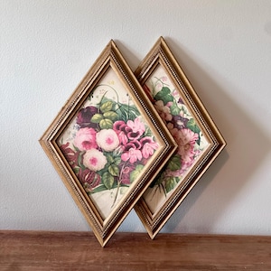 May include: Two framed floral prints. Each print features a bouquet of pink and purple flowers with green leaves. The frames are diamond-shaped with a gold finish and a beaded edge. The prints are displayed on a wooden surface.