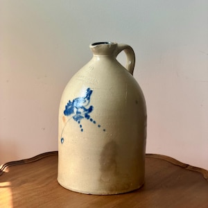 May include: A beige stoneware jug with a single handle and a narrow neck. The body features a blue floral design and the stamped text "COWDEN & WILCOX" above the number "2". The surface shows natural variations in color and texture.