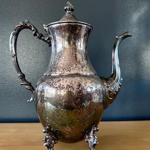 Puede incluir: Una tetera de plata con un diseño ornamentado y tres patas. La tetera tiene un acabado brillante y está ligeramente deslustrado.