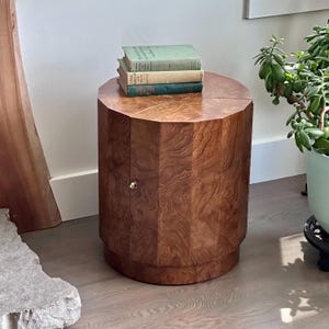 May include: A wooden side table with a geometric design and a warm, brown finish. The table has a small door with a gold-colored knob. Three vintage books in shades of green and tan rest on top of the table. The table is approximately 24 inches tall.