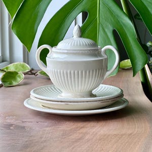 Może przedstawiać: Kremowa cukiernica ceramiczna z pokrywką i dwoma pasującymi spodkami. Cukiernica ma żebrowany wzór i dwa zakrzywione uchwyty. Spodki są ułożone w stos i mają podobny żebrowany wzór. Zestaw stoi na drewnianej powierzchni z zielonymi liśćmi.