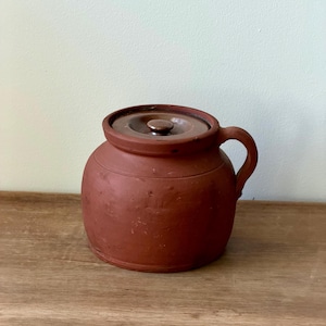 May include: A round, reddish-brown ceramic jar with a lid and a handle. The jar has a slightly textured surface and a dark brown lid with a small knob. The jar is sitting on a wooden surface.