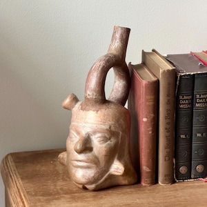May include: A terracotta-colored ceramic vessel shaped like a human head with a stirrup-style spout extending from the top. The sculpture features detailed facial features including eyes, a nose, and a mouth, with a smooth, matte, earthy finish.