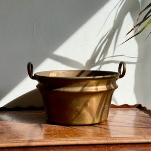 Peut inclure: Une bassine ronde en cuivre métallique dotée d'une large ouverture et de deux poignées circulaires fixées sur les côtés opposés. La surface extérieure présente une teinte chaude et brunie avec des variations de couleur.