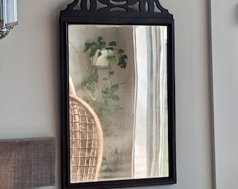 May include: A rectangular, dark-framed mirror with a decorative top detail. The mirror reflects a room with a hanging plant, a woven chair, and a window with sheer curtains. The mirror is mounted on a light-colored wall.