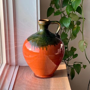 Puede incluir: Jarra de cerámica hecha a mano con asa. La jarra presenta un acabado brillante con una combinación de colores vibrantes. La parte superior es de color verde oscuro, que pasa a un cuerpo naranja brillante. La jarra mide unos 25 cm de altura.