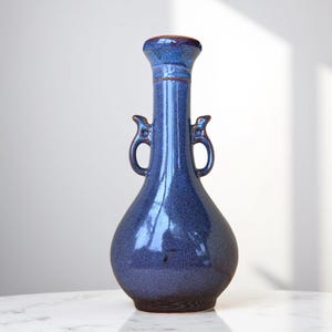 May include: A blue ceramic vase with two handles and a textured glaze. The vase has a round body and a tall, narrow neck.