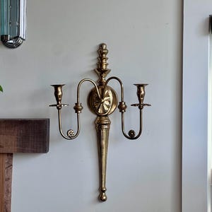 May include: A brass wall sconce with two candle holders. The sconce has a decorative design with a central circular element and a long, tapered base. The candle holders are curved and adorned with small, round details. The sconce is mounted on a white wall.