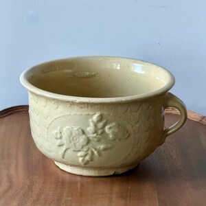 May include: A pale yellow ceramic chamber pot featuring a raised floral relief design on the front and a textured scale-like pattern around the exterior. The vessel includes a single side handle and a wide, circular opening with a smooth rim.