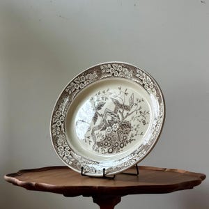 May include: A round ceramic plate featuring a cream-colored center with a brown transferware pattern of stylized floral branches and geometric ribbons. The wide rim is decorated with a detailed border of flowers and intricate scrollwork.