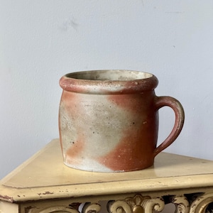 May include: A cylindrical ceramic mug featuring a mottled, earthy color palette of muted terracotta orange and weathered off-white. The vessel has a thick, rounded rim, a smooth exterior surface with visible speckling, and a single curved handle.