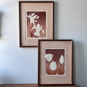 May include: Two framed floral art prints. The prints feature white textured flowers against a dark red-brown background. The frames are a dark brown color, and the mats are a light pink. The top print shows irises, and the bottom print shows tulips.