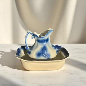 Könnte beinhalten: Antikes cremefarbenes Waschbecken-Set mit blau-weißem Blumendesign. Das Set besteht aus einem Krug und einem Becken, beide mit aufwendigen Mustern. Der Krug hat einen gebogenen Griff und einen Ausguss. Das Becken ist oval geformt.